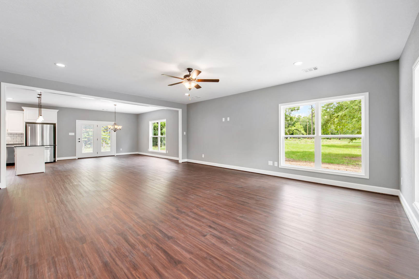 Spacious room with hardwood flooring, ceiling fan with light fixture, double doors, and windows overlooking grassy field and trees
