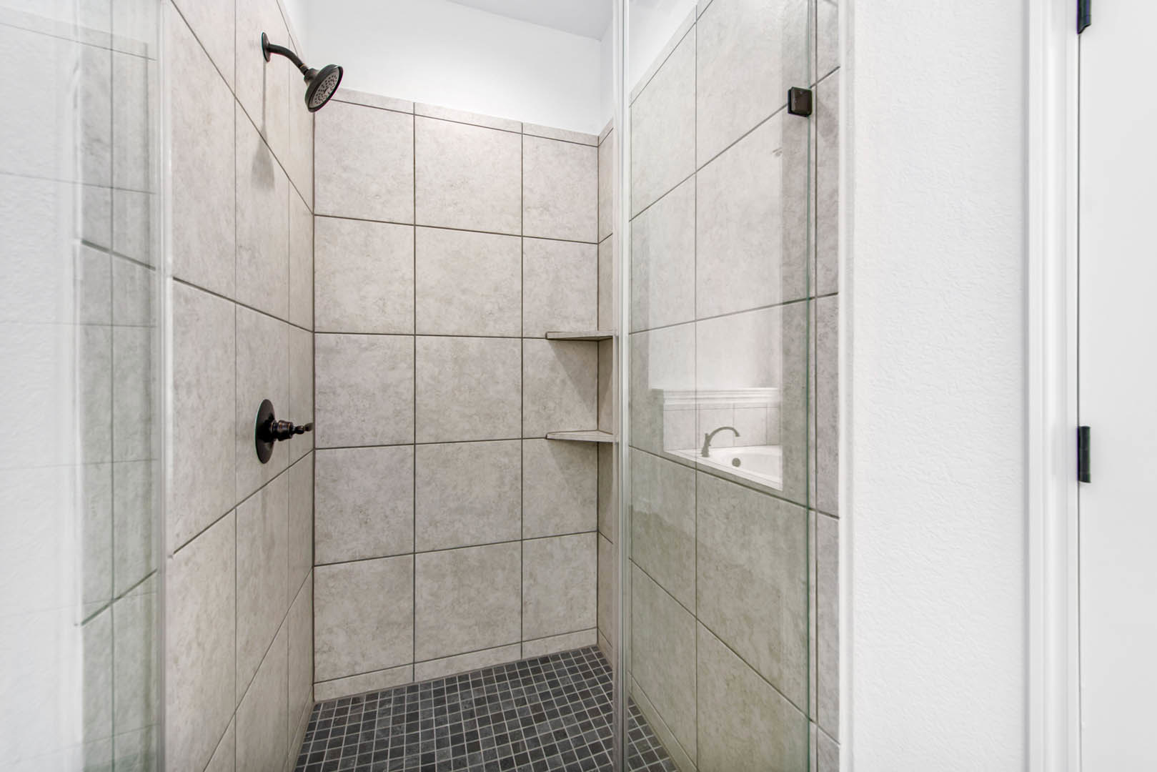Frameless glass shower door with chrome handle, white tile walls, built-in shower head, and light gray tile flooring in a modern bathroom