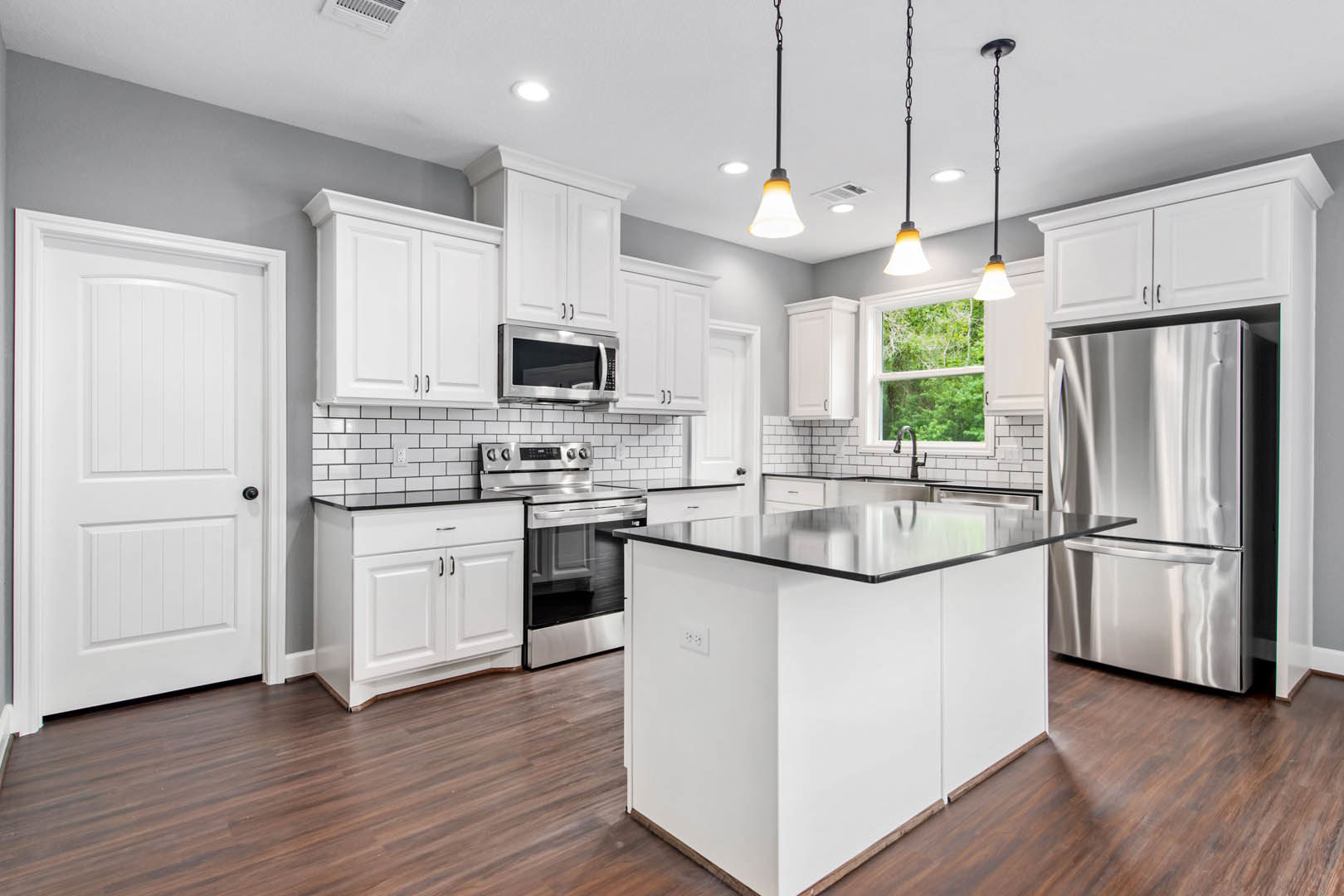 White kitchen with shaker cabinets, stainless steel refrigerator and oven, black countertop island, open microwave door, white door with black knob, light wood flooring