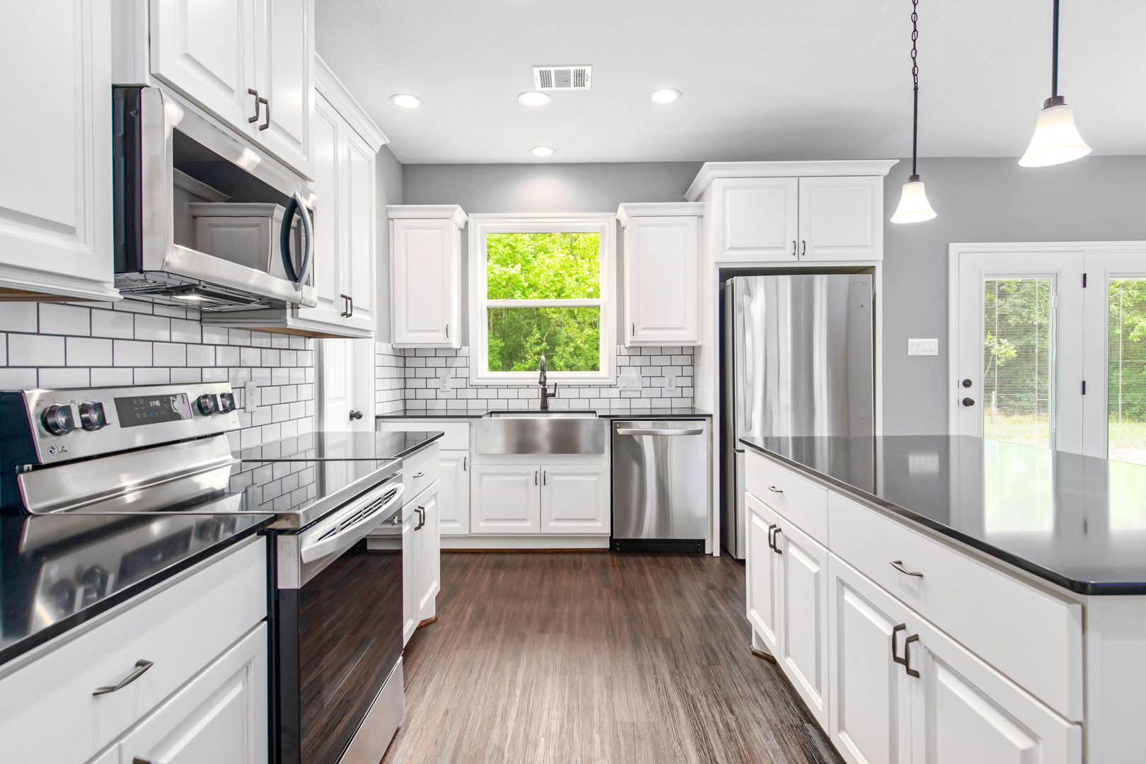 White kitchen cabinets, stainless steel refrigerator and oven, wood flooring, quartz countertops, window overlooking trees, pendant light fixture