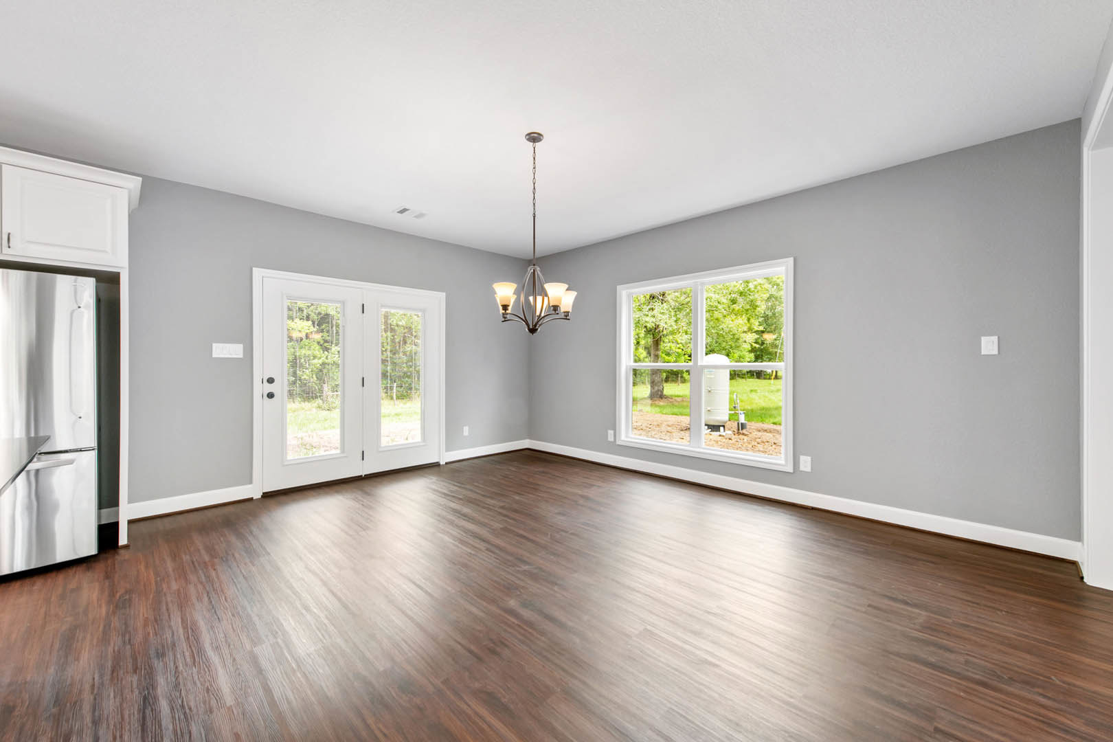 Open living space with hardwood flooring, ornate chandelier, white double doors with glass panels, large window, and partial view of a countertop
