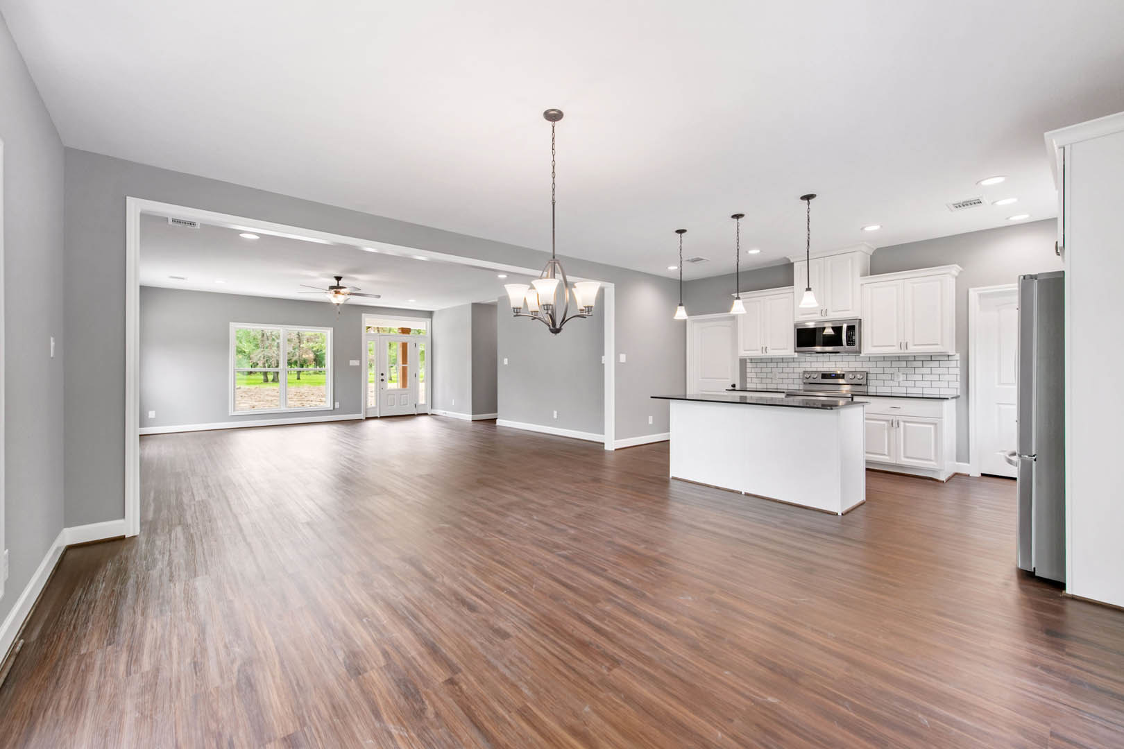 Spacious open floor plan featuring a central kitchen island, hardwood flooring, white walls, recessed lighting, and large windows overlooking a green lawn.