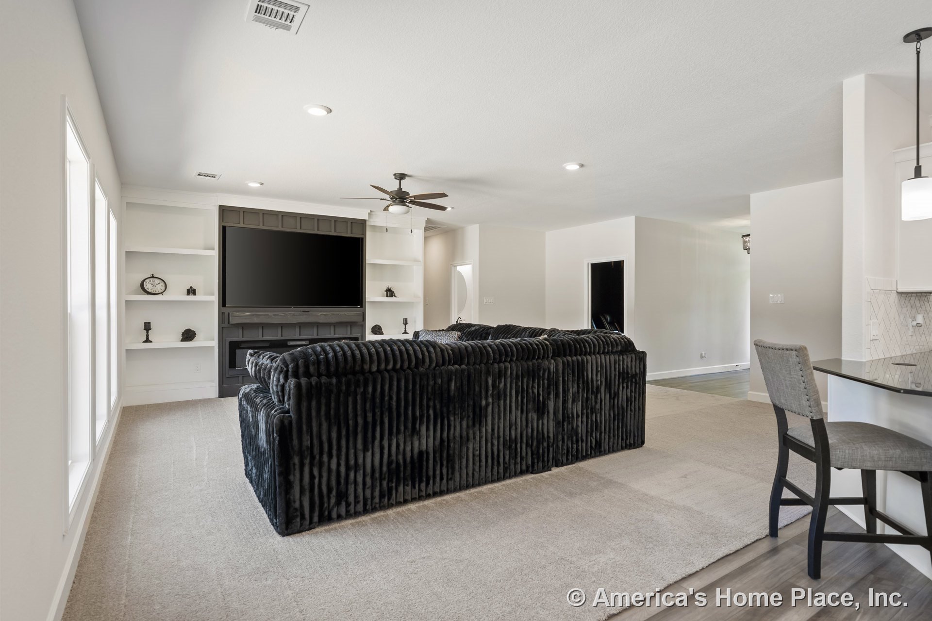 Built-in white shelving flanks a dark entertainment center with integrated fireplace, recessed ceiling lights, carpeted floor, ceiling fan with light, and large windows providing