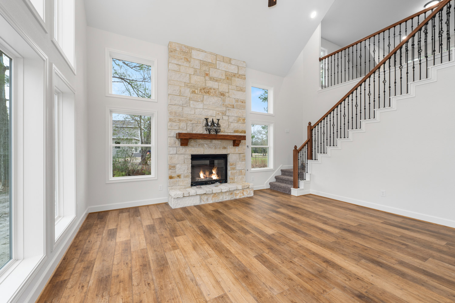 Open living area featuring a modern fireplace with active flames, hardwood floors, and a staircase with metal railings; large windows reveal tree branches and a neighboring house.
