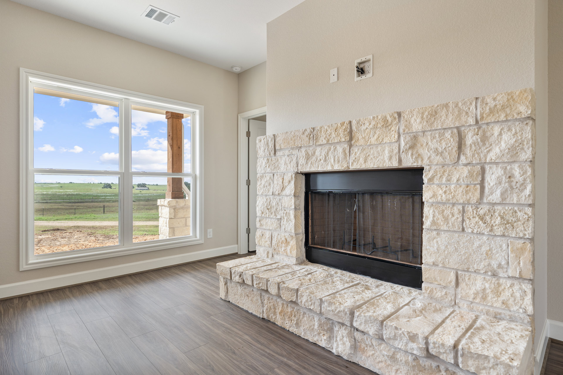 Stone fireplace with black frame set in a living room, wooden post nearby, large window overlooking grassy field, wall outlet visible, stone wall and door frame in background