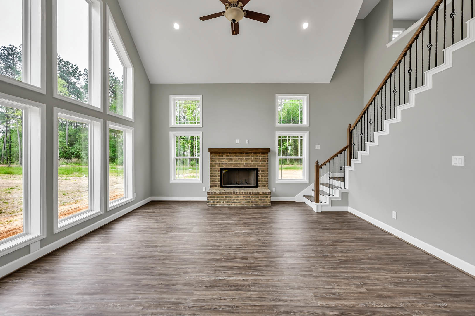 Brick fireplace with wood mantel, hardwood floors, staircase with wooden railing, ceiling fan with light, window framed by molding, brick accent wall featuring a yellow sign.