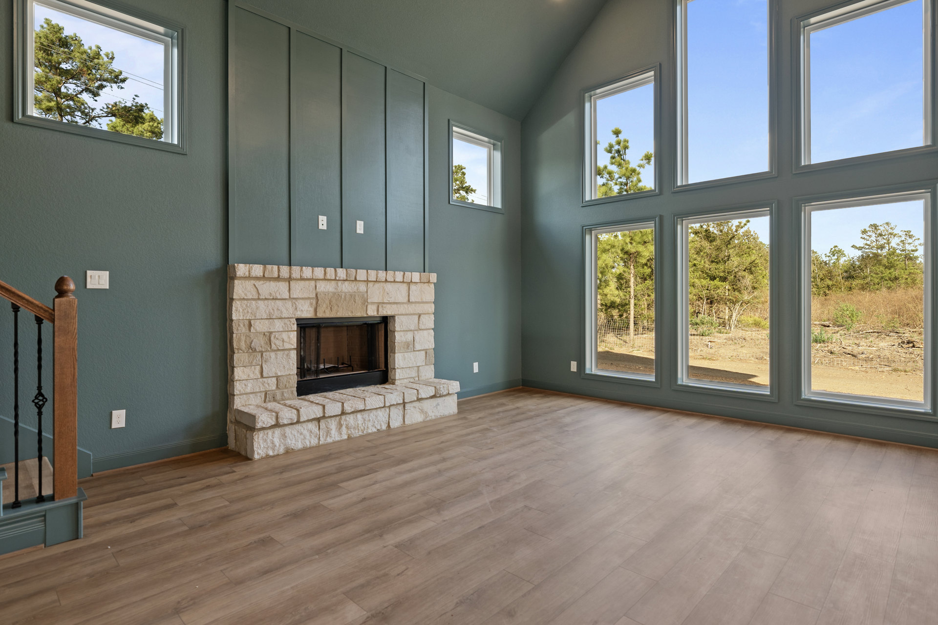 Brick fireplace with black mesh door, wood flooring, metal railing, large windows framing outdoor trees