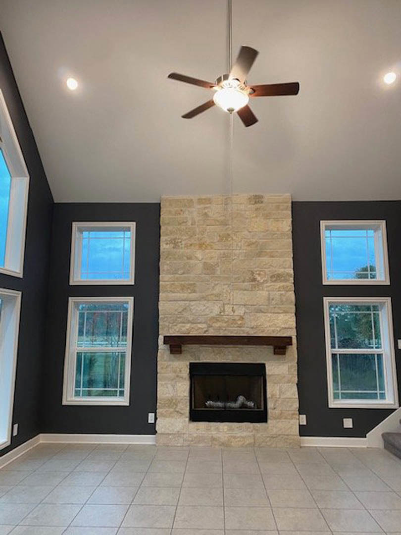 Ceiling fan with light fixture above a tiled floor in a living room featuring a brick fireplace, white walls, and large windows.