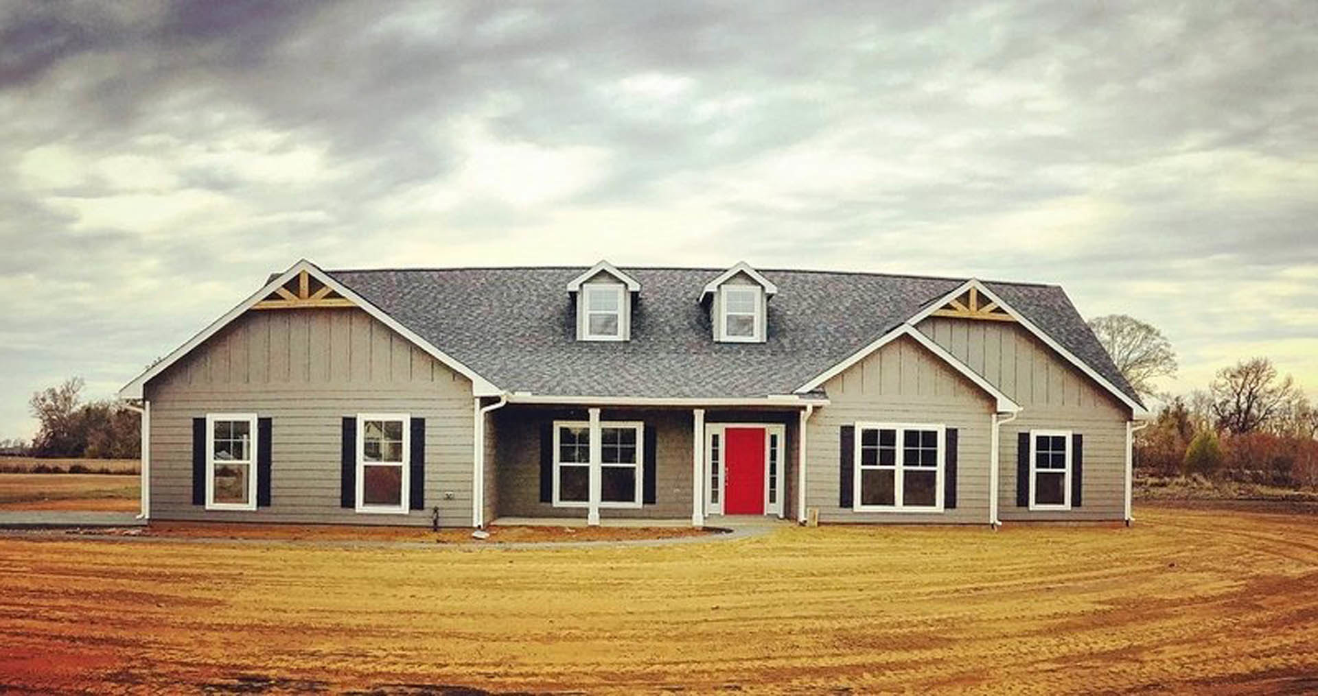 Red front door with white trim, white-framed windows, gray shingle roof, light-colored siding, and landscaped entryway under a partly cloudy sky.