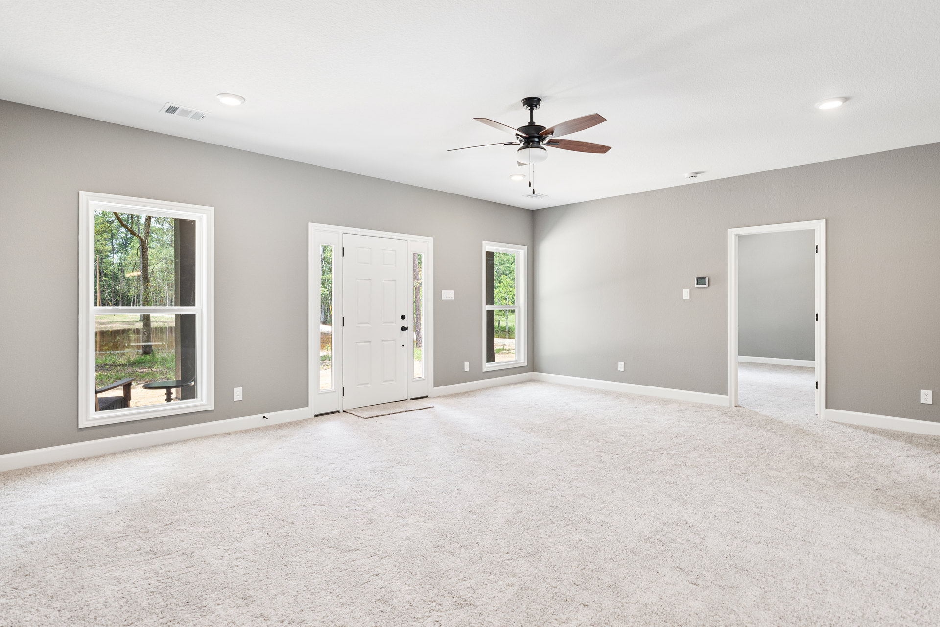 Ceiling fan with light fixture above white carpeted floor, large windows showing tree and water view, white walls, glass-paneled door