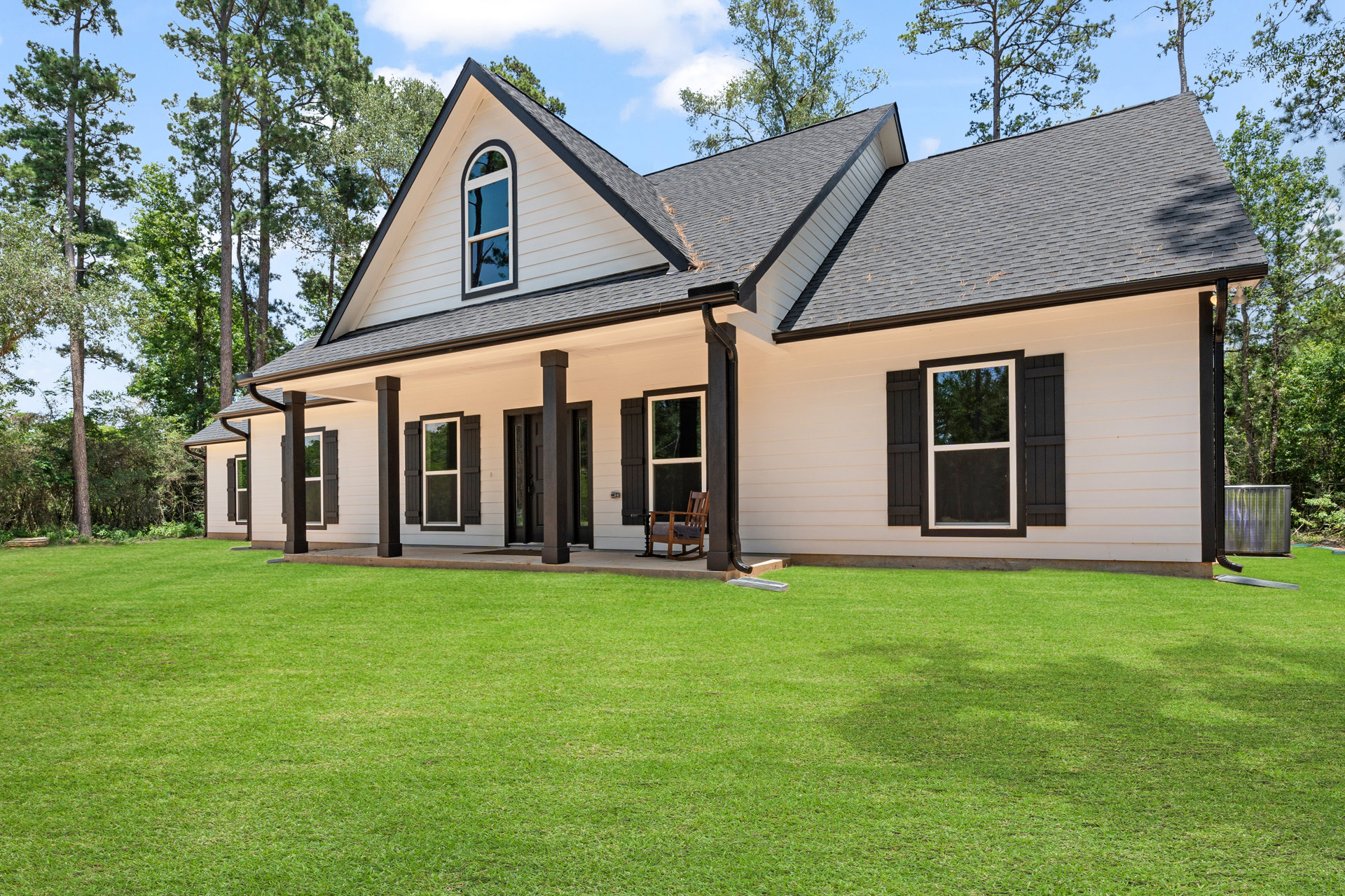 White siding house with black front door, wooden rocking chair on porch, large windows reflecting nearby trees, green lawn bordered by mature trees, metal grill in foreground.