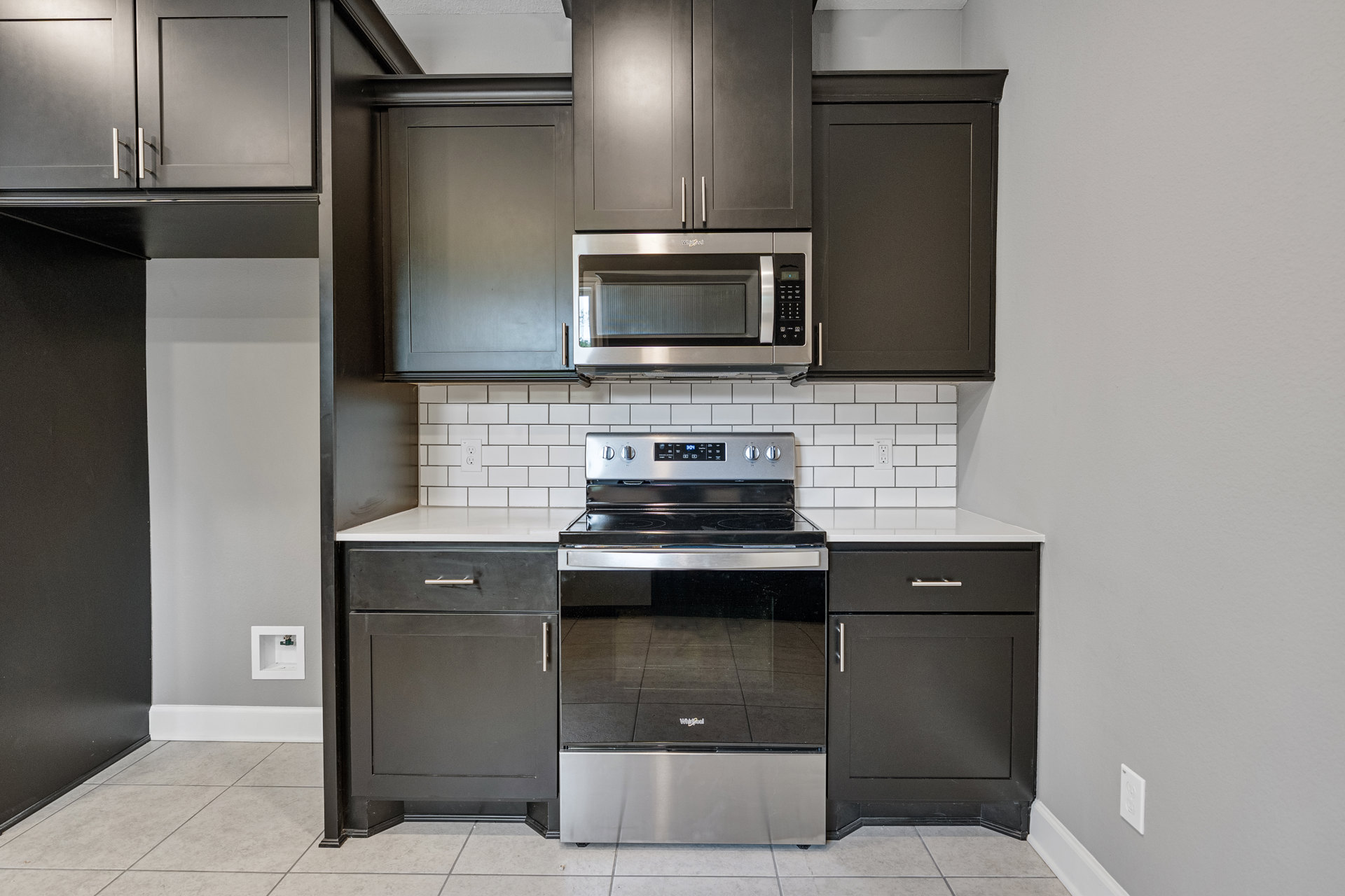 Black kitchen cabinets with silver handles, stainless steel stove and microwave, speckled black countertop, grey cabinet doors, and a door with a metal handle.