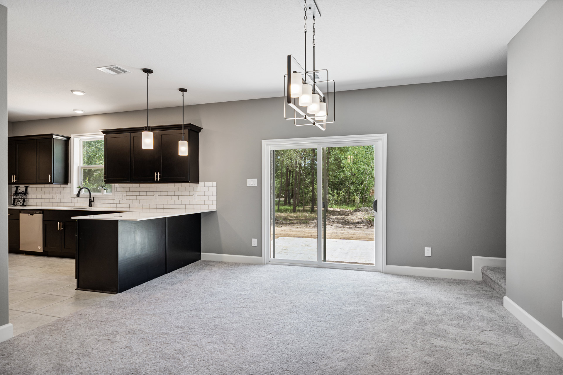 Open kitchen with black cabinets and silver handles, white ceiling lights, light wood flooring, and a sliding glass door overlooking a forest.