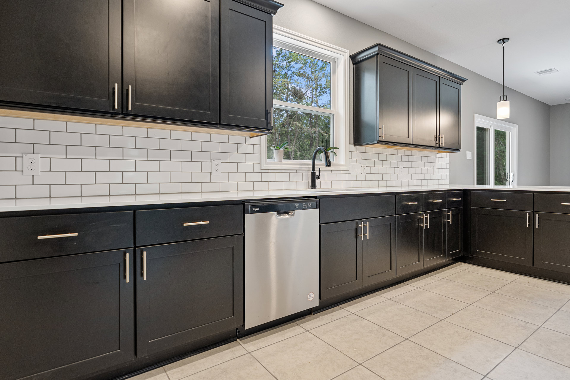 Black kitchen cabinets with silver refrigerator, white tile floor, window overlooking trees, and stainless steel sink