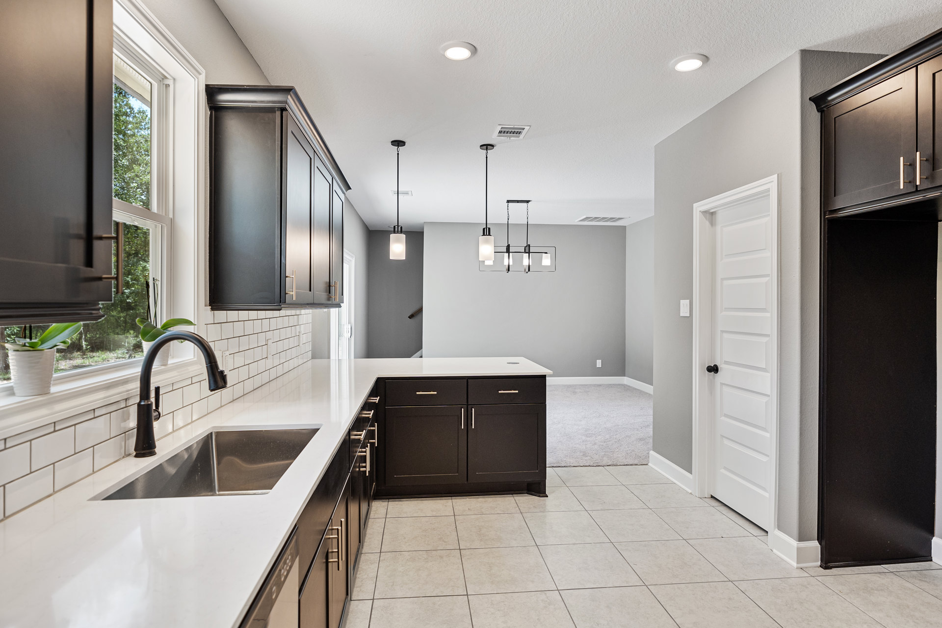 Modern kitchen with matte black lower cabinets, white quartz countertop, stainless steel undermount sink, chrome faucet, small potted plant, and white door with black hardware