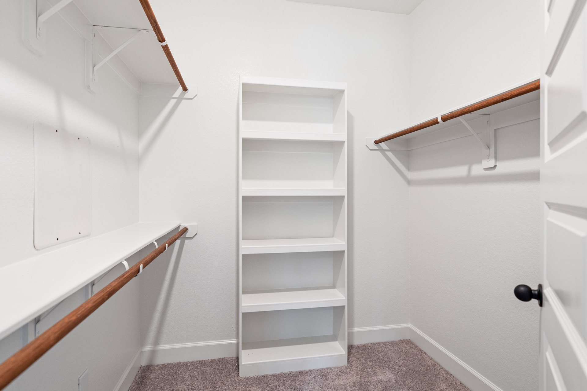 White built-in shelves with wooden accent bar mounted on a plaster wall, black door knob visible nearby, carpeted floor in a bright white room.