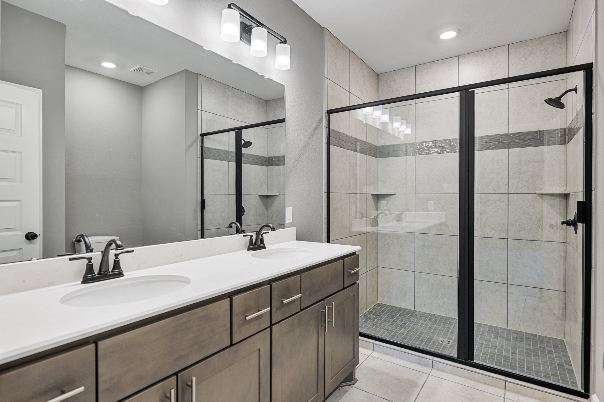 Bathroom with glass-enclosed shower, double vanity with white sinks and chrome faucets, large mirror above countertop, light tile flooring, and white cabinetry with black handles
