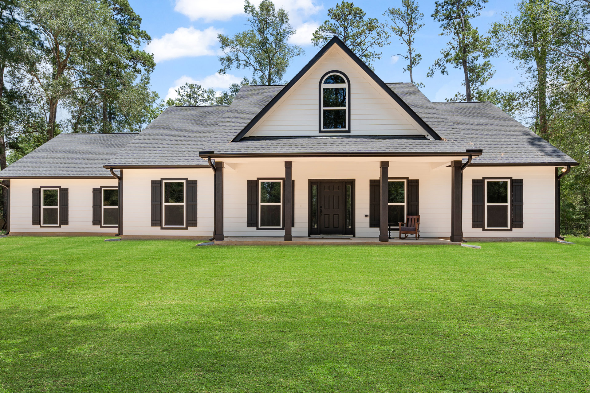 Two-story white farmhouse with black-framed windows, black front door with glass panels, wooden rocking chair on covered porch, green lawn in foreground, trees and cloudy sky in