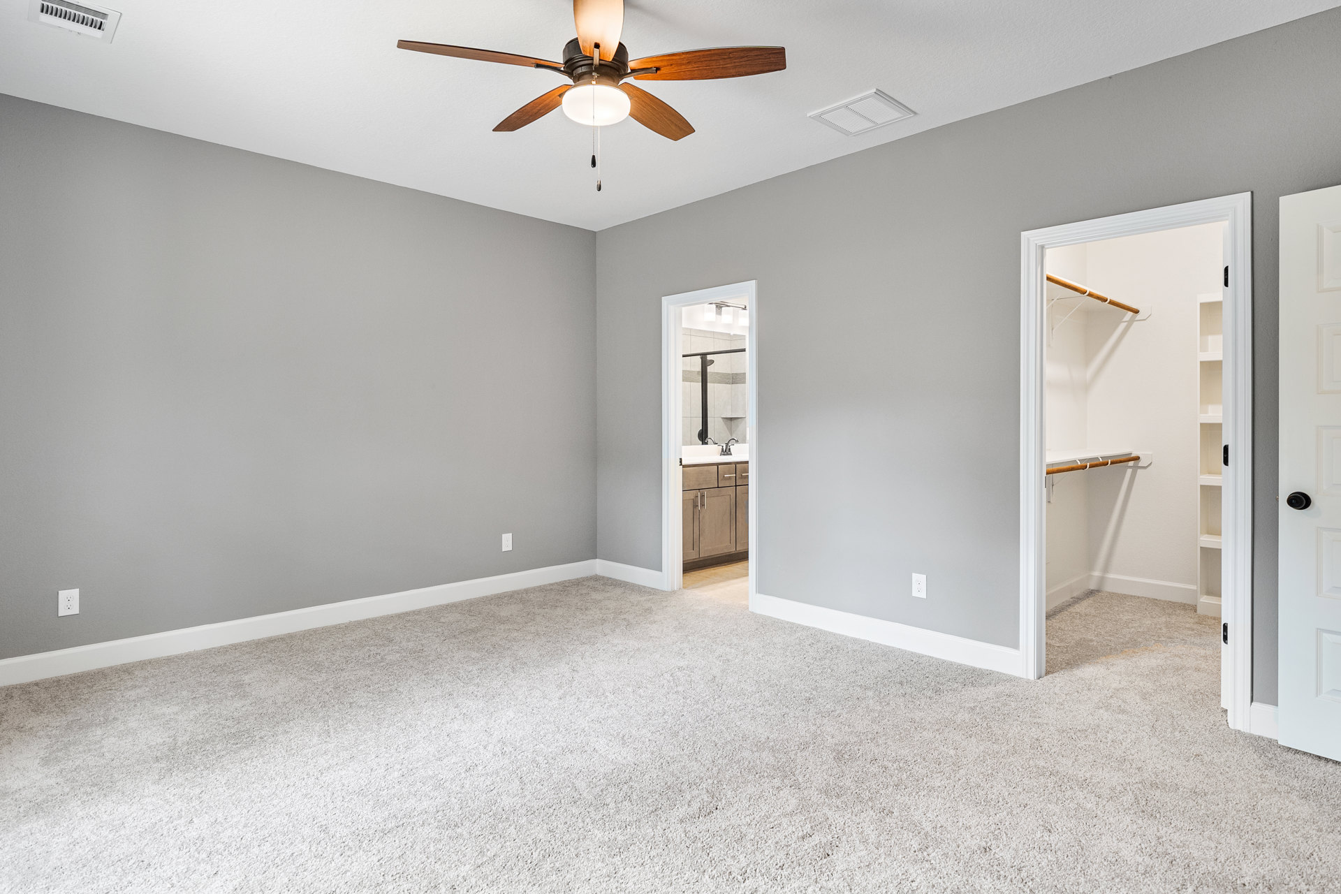 Ceiling fan with light fixture above white carpeted floor, built-in sink with kitchen cabinet, white vent on ceiling, door leading to adjacent room, neutral wall finishes.