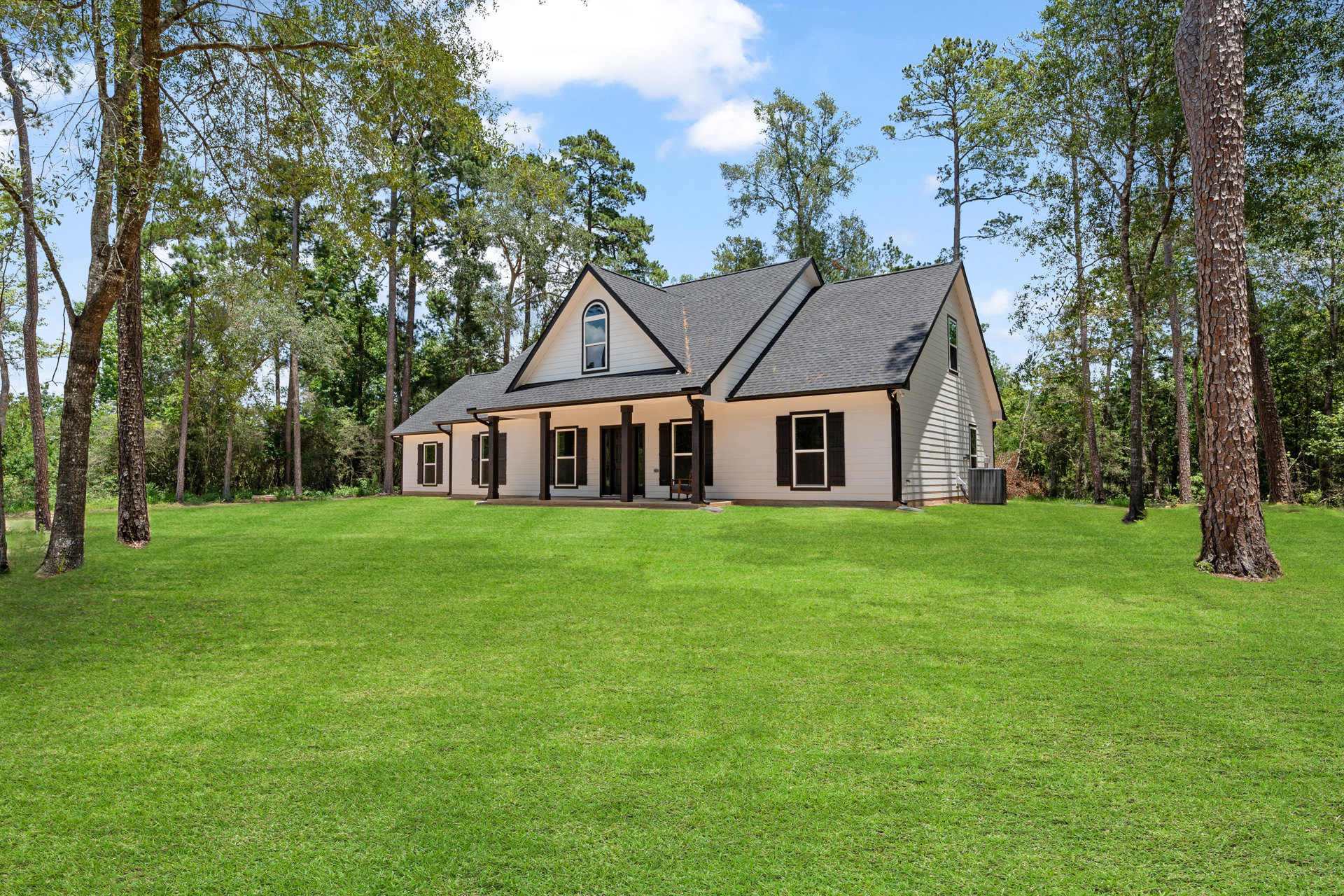 Two-story farmhouse with white siding, dark roof, covered front porch, expansive green lawn, mature trees in background, large windows with white frames, partly cloudy sky overhead