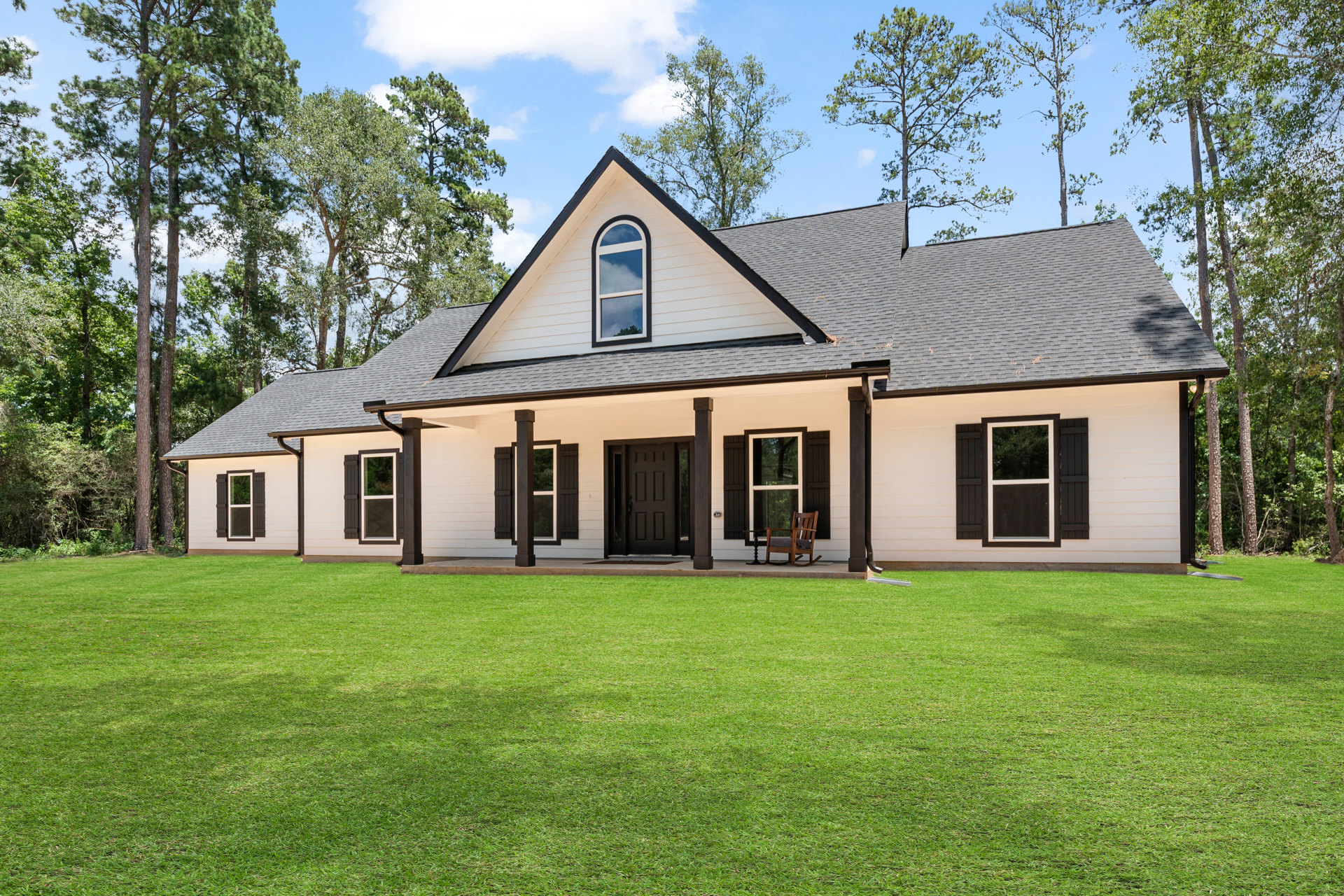 White house with black roof, white-framed windows reflecting blue sky, wooden rocking chair on porch, green lawn in foreground, Little White House visible in background.