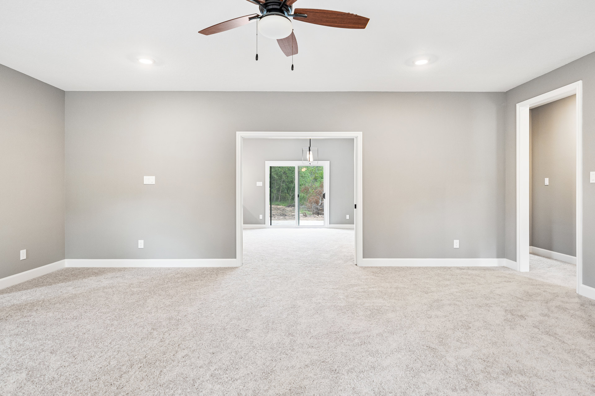 Ceiling fan with light fixture mounted on white ceiling, sliding glass door revealing trees outside, white carpeted floor, two interior doors, light switch near doorway