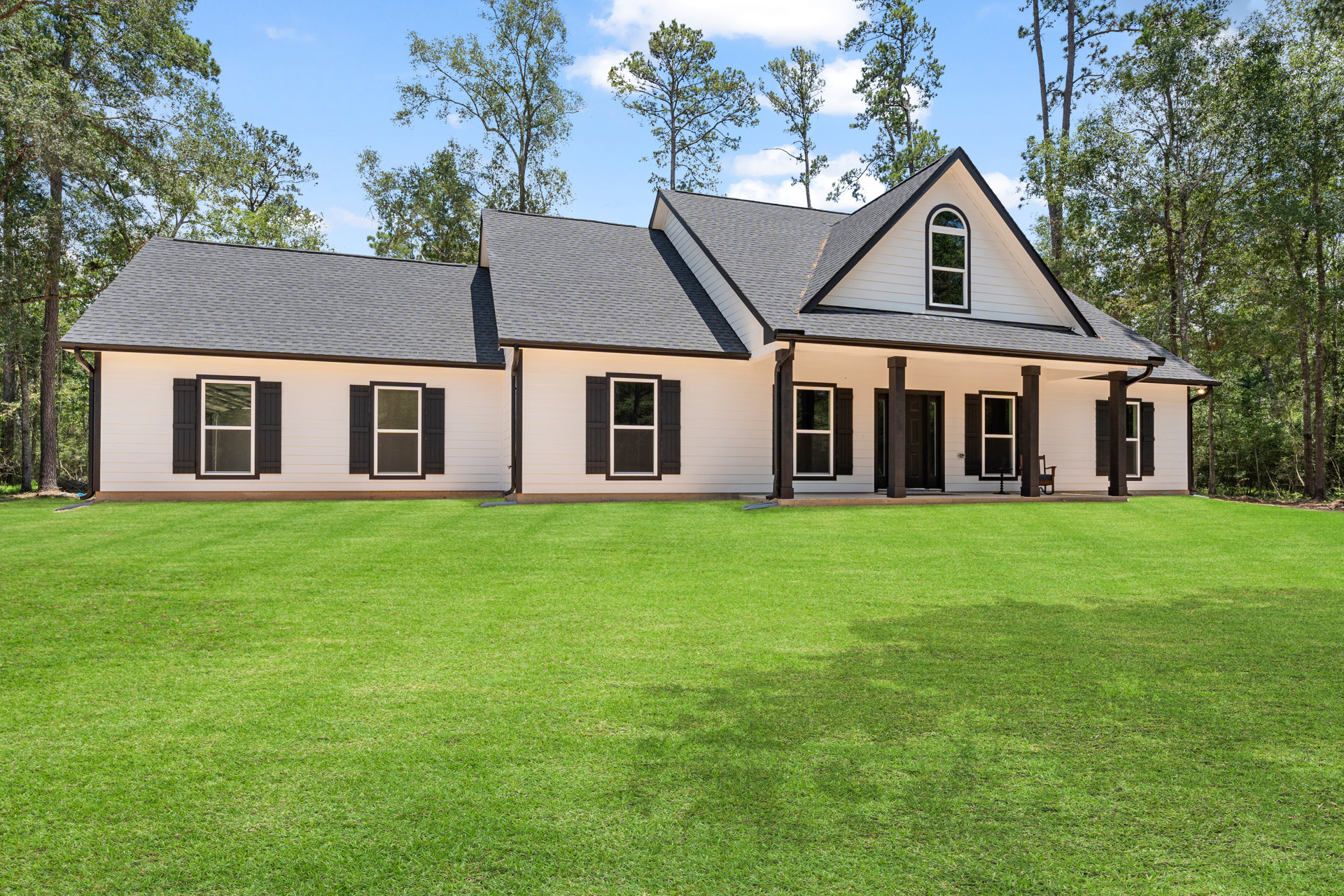 White farmhouse with black trim, large windows, manicured green lawn, mature trees behind the roof, clear blue sky