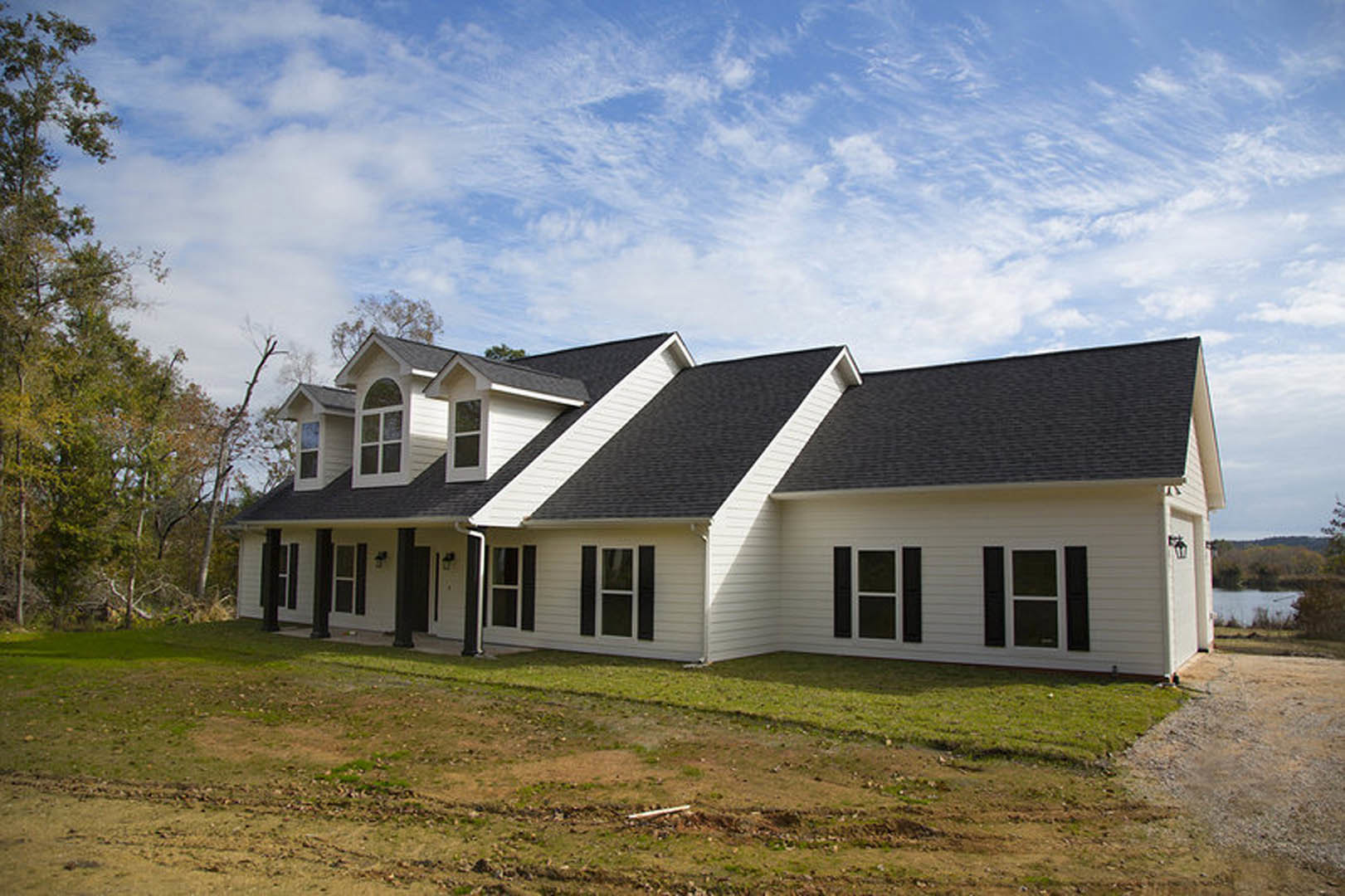 Two-story house with black roof, white-framed windows, beige siding, green lawn, dirt path, and blue sky with scattered clouds