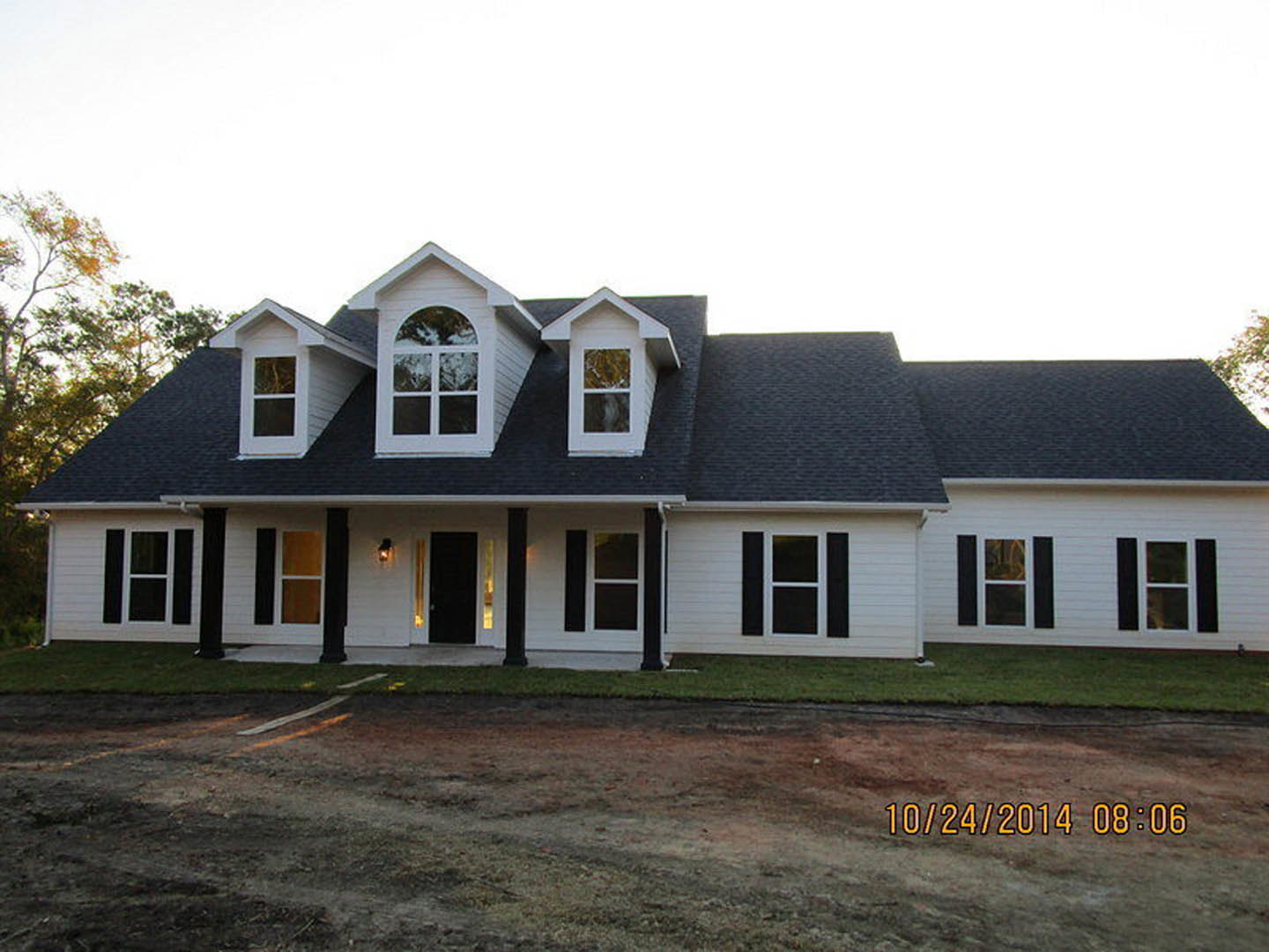 White two-story house with black trim, dormer window, black front door with white trim, covered porch, manicured lawn, and mature trees in the background
