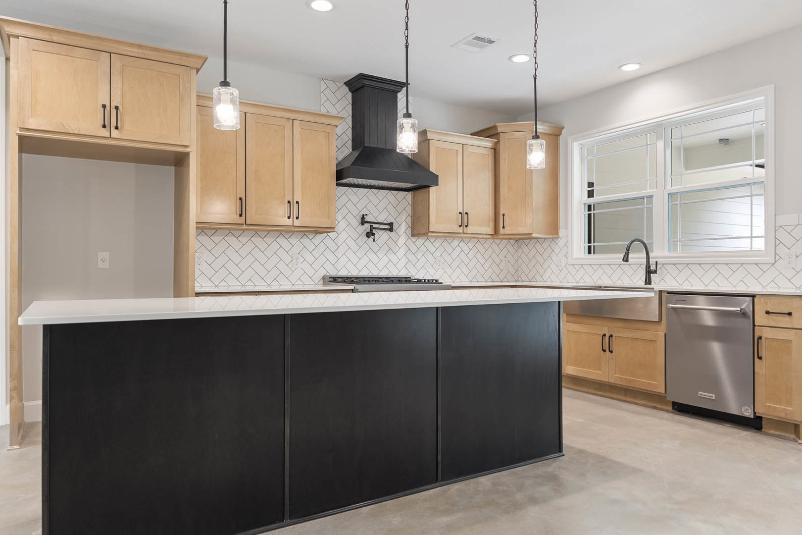 Black kitchen island with white countertop, light wood cabinets, stainless steel dishwasher with handle, glass jar on counter, modern pendant light, large window above sink.