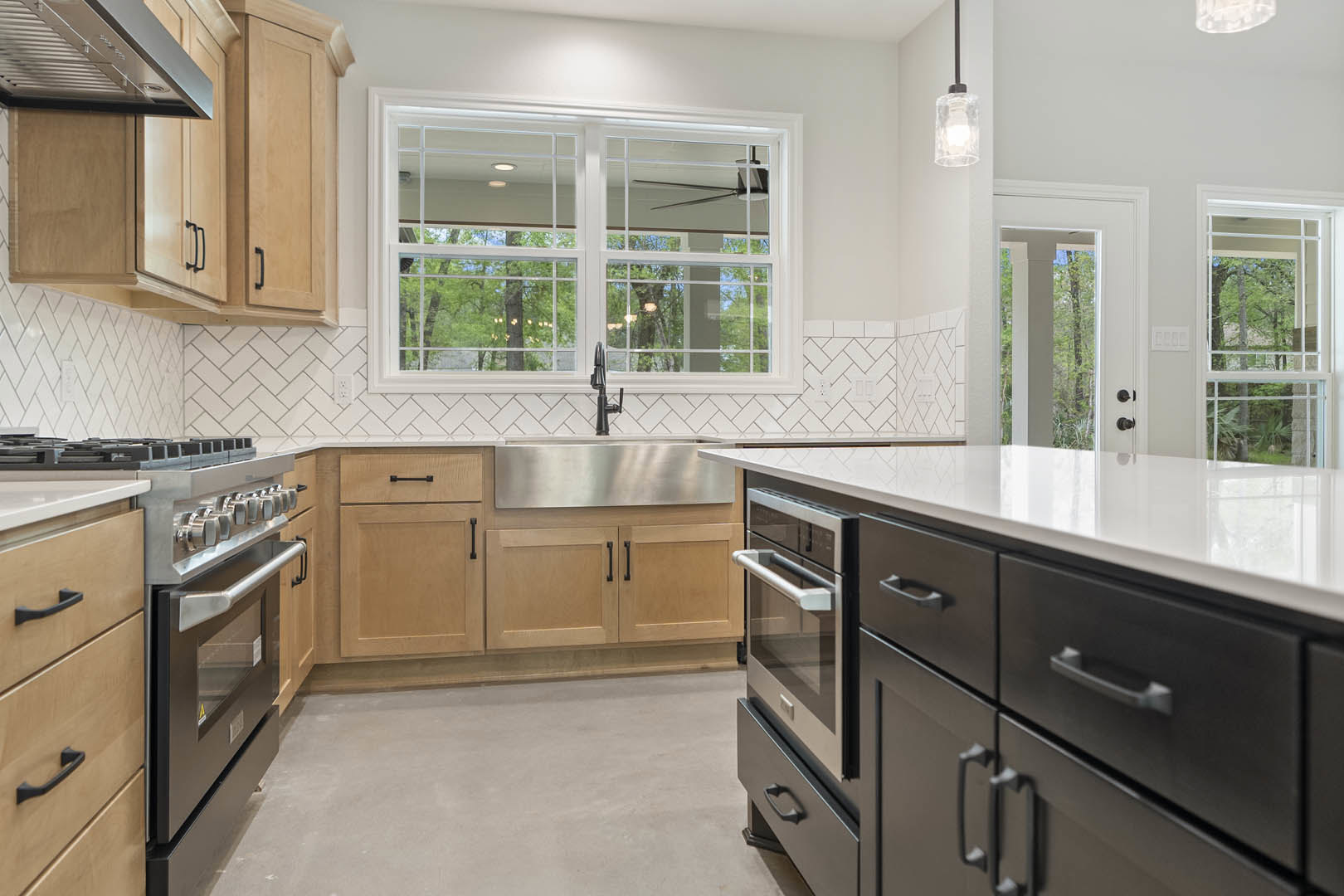 Modern kitchen with white cabinets, stainless steel sink, stone countertop, large window with fan, concrete floor, built-in oven, and open drawer displaying a plastic container
