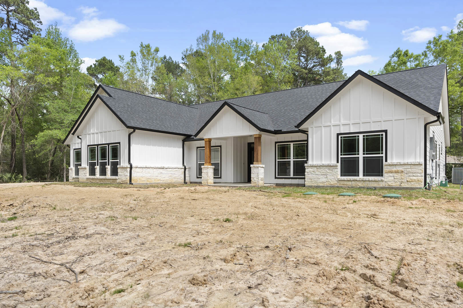 Modern house with black roof and white-framed windows, surrounded by a dirt field and leafless tree under cloudy sky