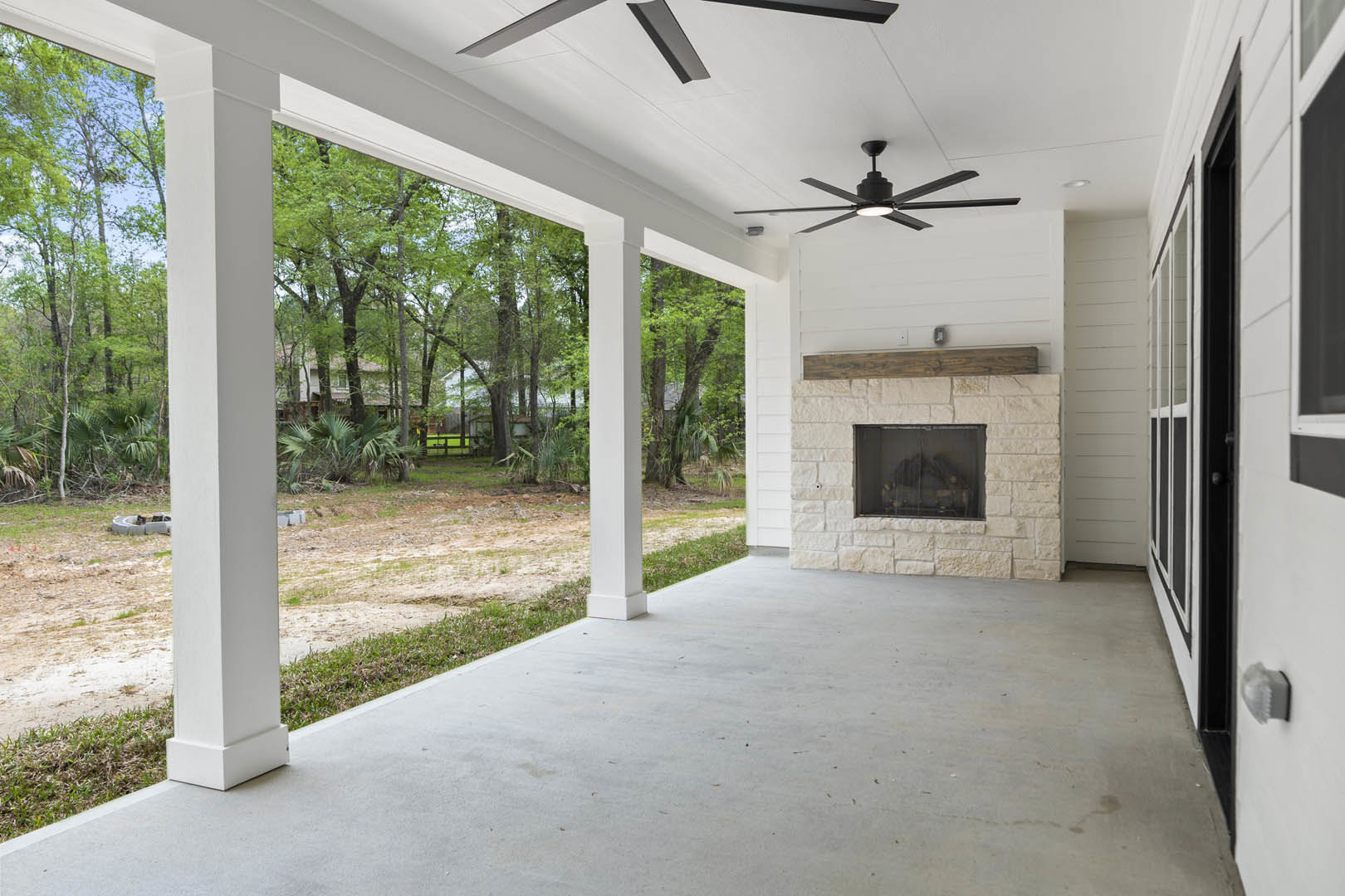 Spacious white living room featuring a wood-burning fireplace, ceiling fan on a white ceiling, large window with glass pane, and concrete walkway with pillars visible outside.