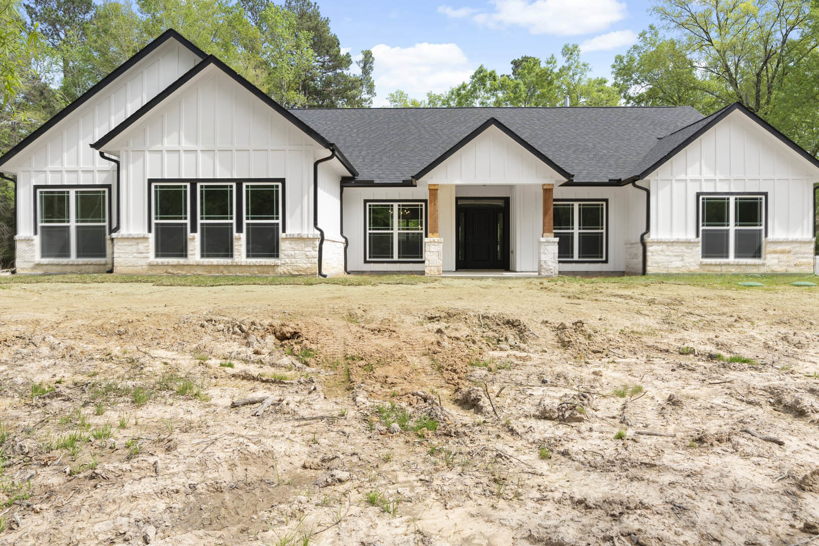 White house with black roof and black front door, surrounded by a dirt patch with sparse grass, large windows, and cloudy sky overhead