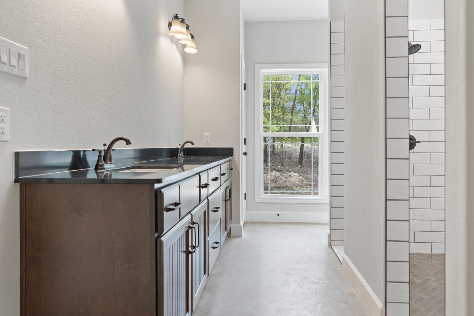 Bathroom with large window overlooking trees, white tile floor, open door, wall-mounted row of lights, modern sink with cabinetry and countertop, close-up of light switch.