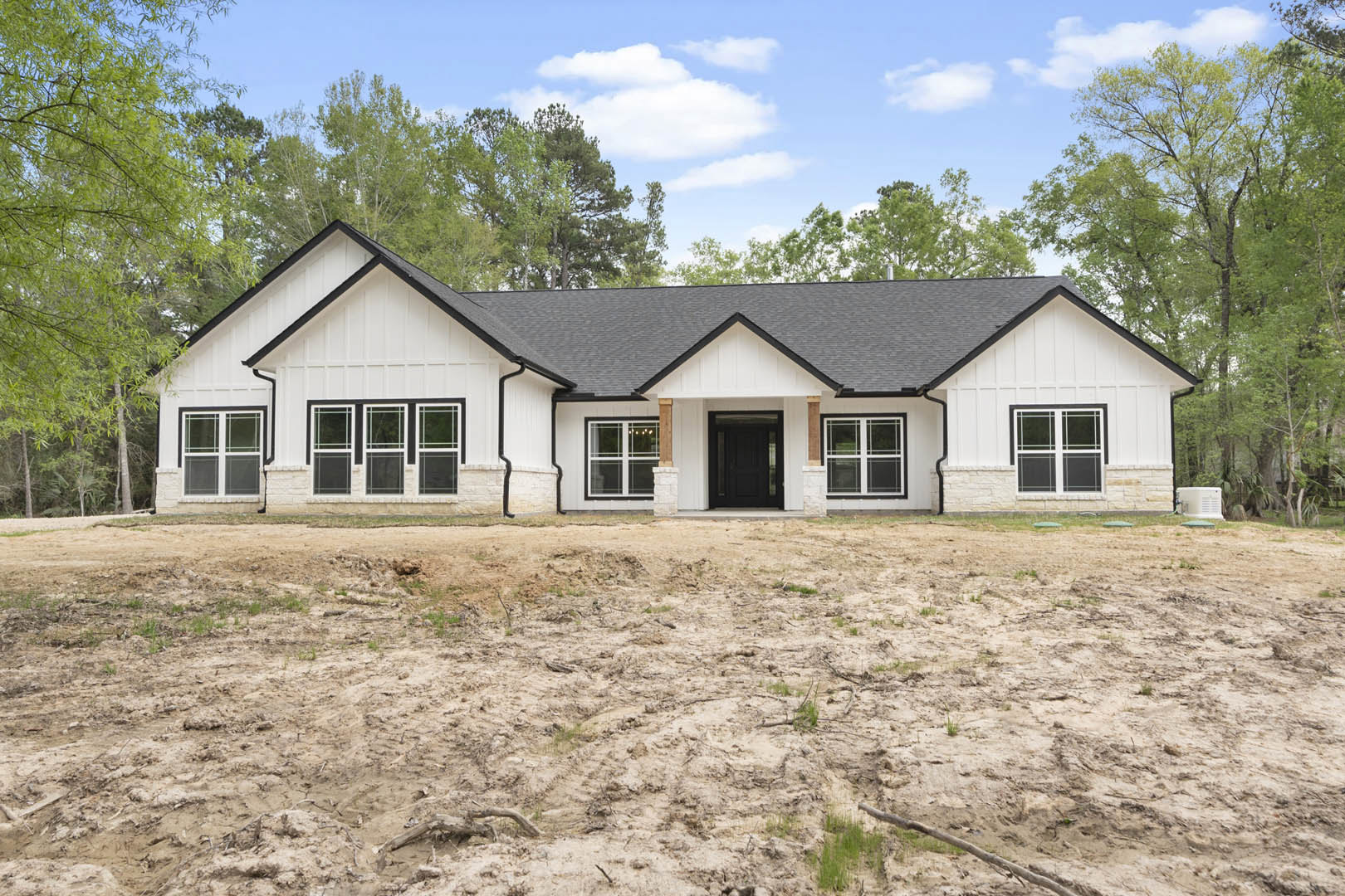 Modern white house with black-framed windows and a black glass-paneled front door, surrounded by a dirt field and sparse landscaping under a cloudy sky