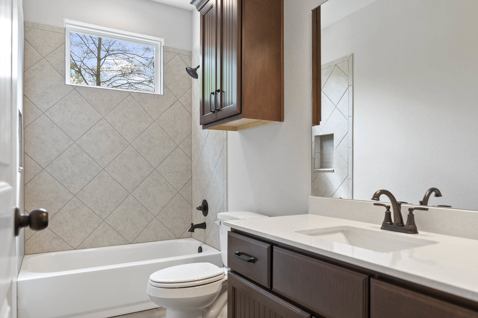 Bathroom with white toilet, modern sink and cabinet, tiled walls, window overlooking a tree, shower head with hose, neutral finishes.