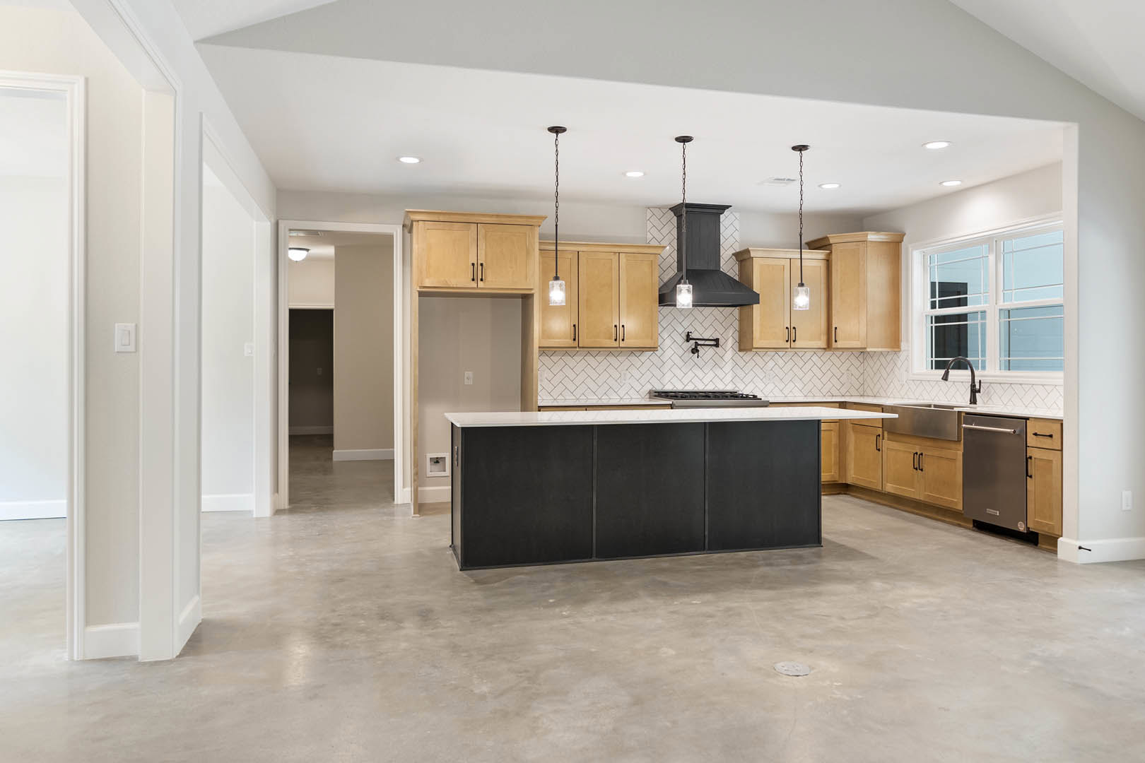 Kitchen featuring a matte black island, light wood cabinets, concrete flooring, white-paned window, black range hood with integrated lighting, and stainless steel dishwasher.