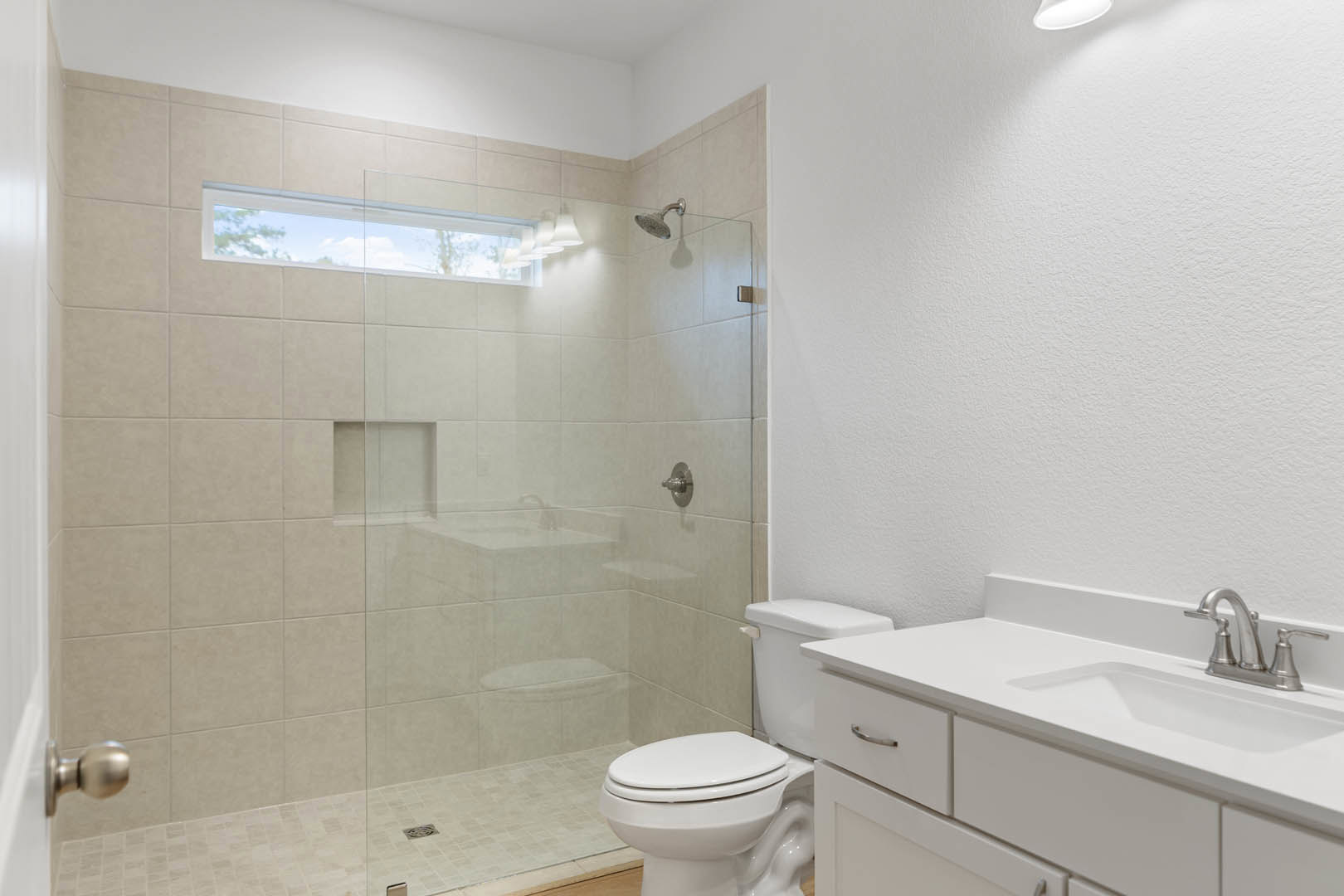 Bathroom with glass shower enclosure, white sink with silver faucet, wall-mounted shower head, toilet beside sink, large window showing blue sky, light-colored tile walls and floor