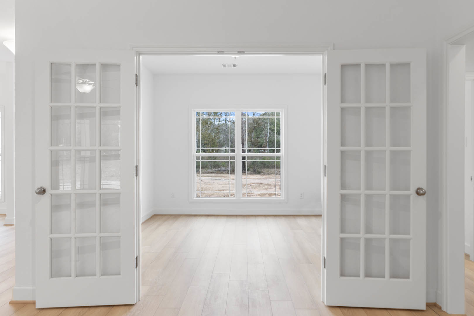 Wood-floored room with white walls, double glass-paneled doors, and a window overlooking a forest