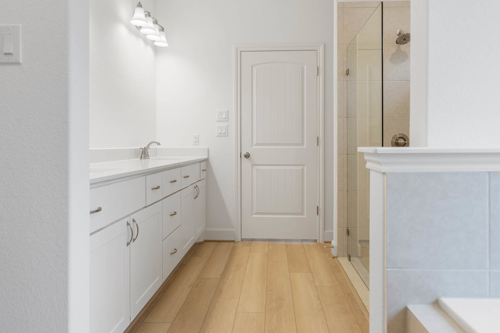 Bathroom with wood flooring, white cabinets and drawers, white countertop with silver faucet, white tile wall, white door with silver knob, row of white lamps above vanity