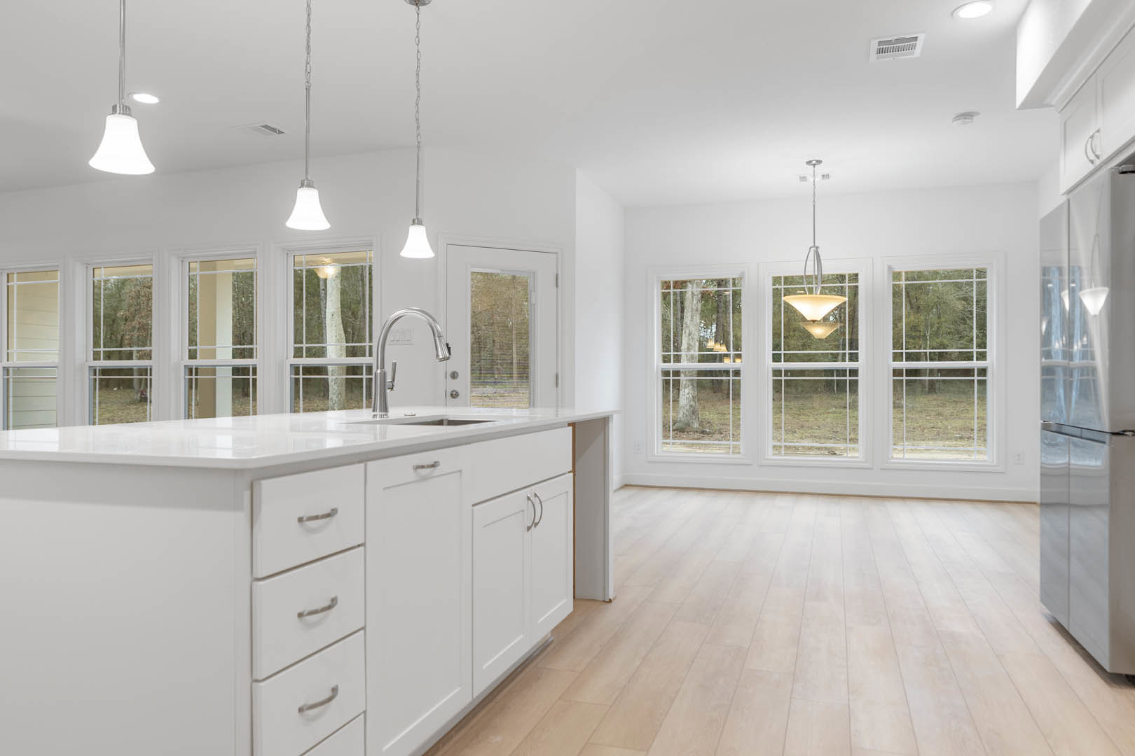 White kitchen featuring white cabinets, wood flooring, stainless steel faucet, undermount sink, drawers, ceiling light fixture, and a window overlooking a forest.
