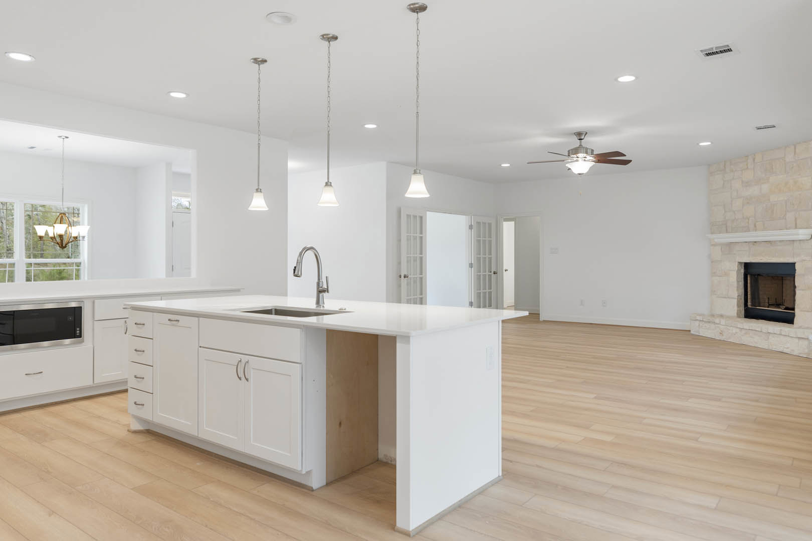 White kitchen island with built-in sink, white cabinetry, stainless steel appliances, ceiling fan, stone fireplace, and hardwood flooring
