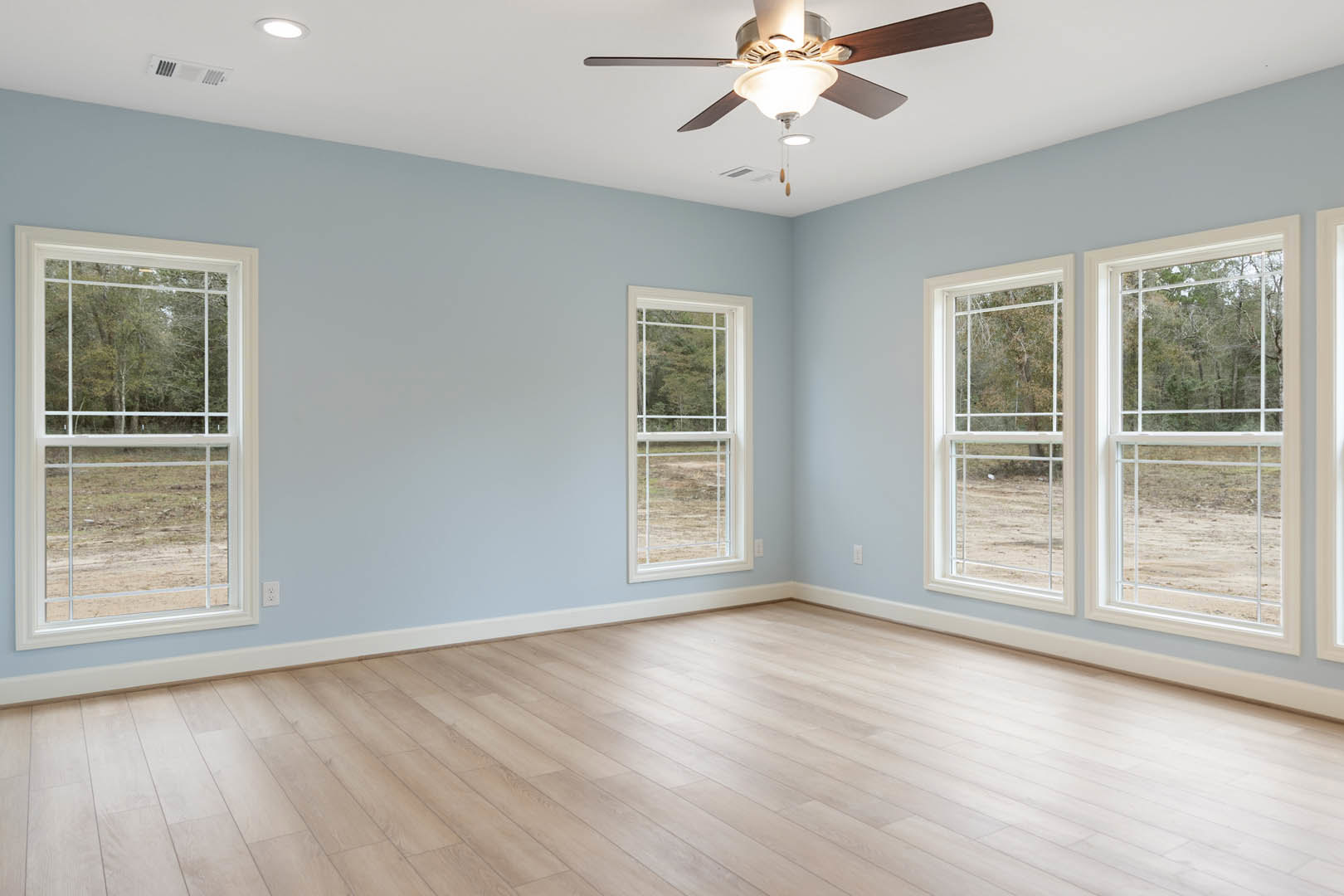 Living room with wood flooring, ceiling fan with light fixture, large windows overlooking trees, white walls, and natural light.