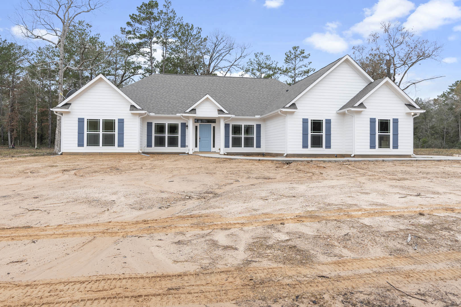 Two-story house with blue front door and white-framed windows, surrounded by dirt yard with tire tracks, gray shingle roof, cloudy sky overhead, and trees in background