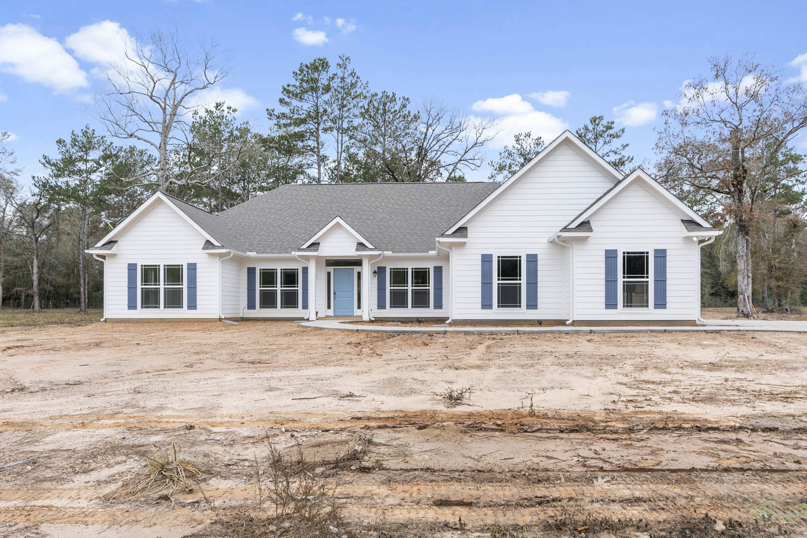 White house with blue doors and windows, surrounded by dirt yard and trees in the background, cloudy sky overhead