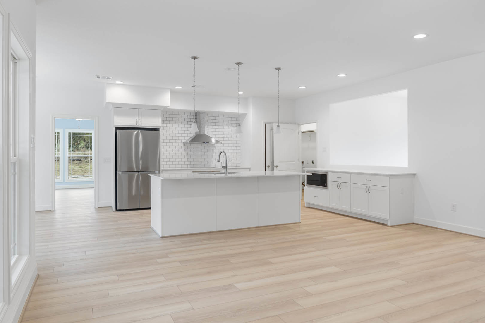 Kitchen with wood flooring, white cabinetry, stainless steel refrigerator, white countertop with built-in sink, white tile backsplash, ceiling light fixture, and window above the