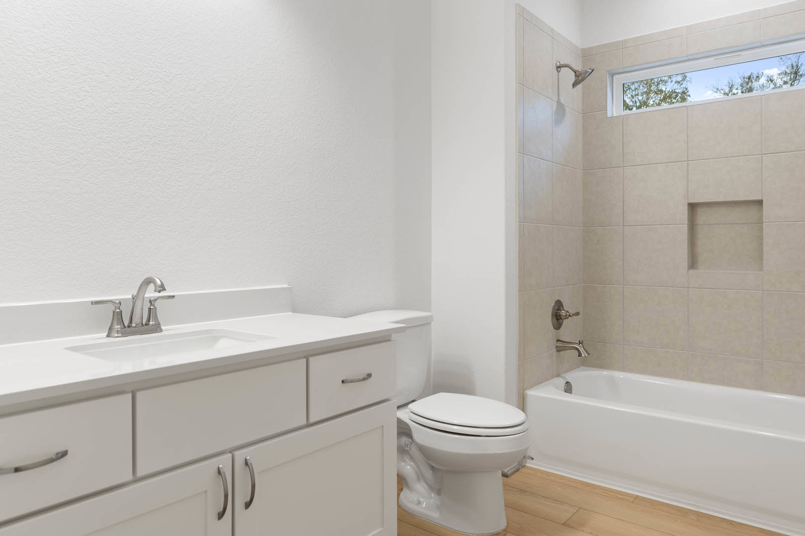 Bathroom with white bathtub and toilet, wood flooring, square-framed artwork on white wall, window showing trees outside, chrome faucet visible.