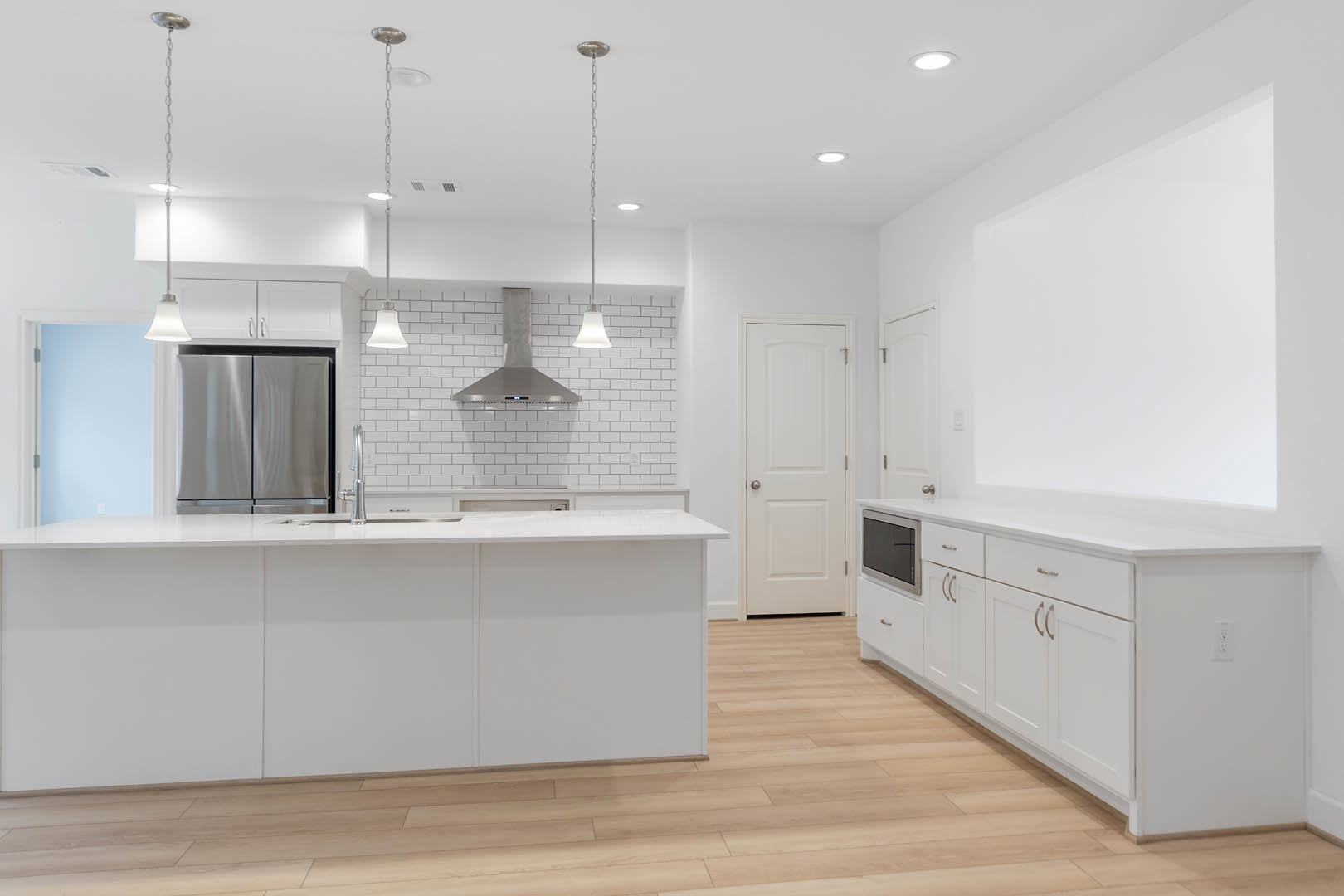 White kitchen with tile floor, white cabinetry, white appliances including refrigerator and microwave, white island with silver faucet, silver door knob, and white light fixture.