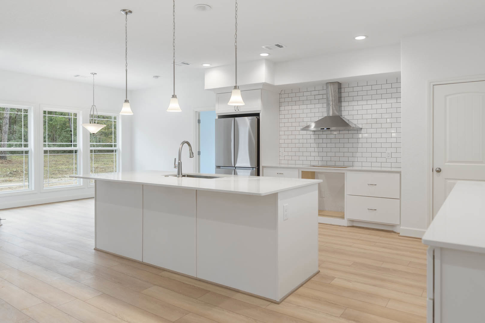 White kitchen with shaker cabinets, black handles, wood plank flooring, white island, stainless steel appliances, undermount sink, tile backsplash, and modern pendant light.